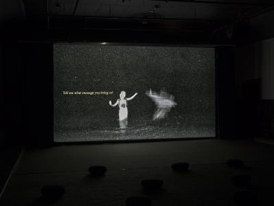 A projected image of two angelic figures. One is blurry, the other in focus. The subjects arms reach for each other atop a staticy gray background. Yellow text reads quote Tell me what message you bring us! 
