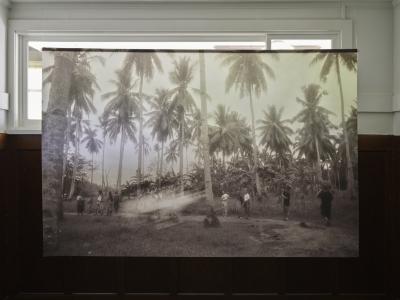 A sepia toned photograph printed on sheer fabric hangs from a dowel attached to the ceiling with fishing wire. The image shows a group of people following a path in a coconut grove, with a ghostly lens flare suggesting the presence of a spirit.