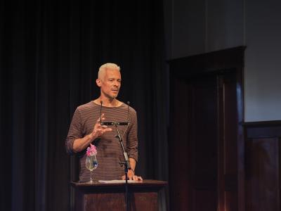 A poet with short white hair and salt-and-pepper stubble addresses a crowd from a lectern. He gestures with his right hand, appearing quite serious and perhaps slightly solemn. He wears  a long-sleeve black and white striped shirt. 