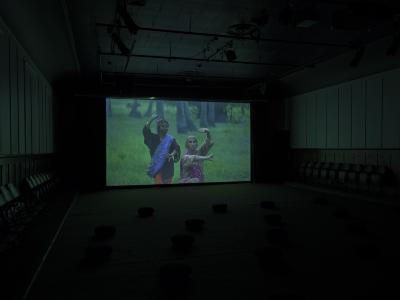 In a projected image, two people with their arms in a dancing motion escapade a lush green forest. Both people are wearing traditional Filipino clothing.