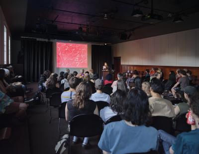 Angled wide-shot of the Grand Luxe Hall. A poet addresses an audience of roughly fifty people from a lectern. They are accompanied by a projection of red and white abstract art displayed to their left. The space has an intimate, sophisticated feel.