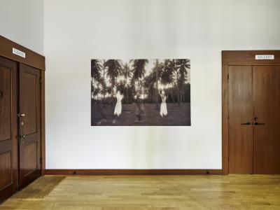 A sepia photograph printed on a textile hangs on a wall between two wooden doors. The image shows a Filipina elder dancing in a coconut grove between two ghostly, iridescent figures.