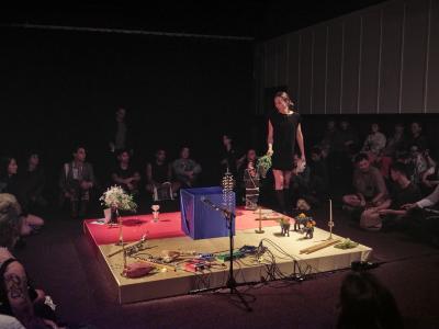 In the Grand Luxe Hall, Bhenji is standing on a square platform holding a bouquet of flowers, surrounded by audience members. The platform is decorated with a red cloth, a blue gift-wrapped box.