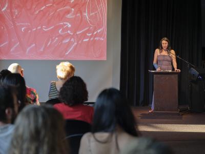 A woman with mid-length brunette hair, a blue cast on her arm, and a mauve sleeveless top addresses a crowd from a lectern. To her right is a projector screen displaying flowing, abstract red and white art. We can see the back of a handful of audience members’ heads, illuminated by sunlight.