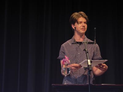 A poet with short brown hair, a mustache, and a short-sleeve, blue and white patterned button down addresses a crowd from a lectern. They hold a small stack of papers with both hands and appear deeply engaged in their speech. 