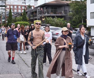 Patrons of the performance walk in a cluster through a back alley. Two people hold small figurines of cows wrapped in palm leaves topped with an orange flower. One person is holding the bamboo buzzer.