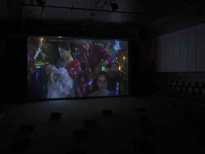An image projected on a screen shows an infant being baptized. A person is having their hair cut by a ritual leader wearing traditional Filipino garments. There are many plants and singular lit candles surrounding the subjects. 