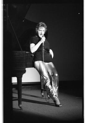 A person leans with their left elbow resting on a grand piano behind them. In their right hand they hold a wired microphone to their mouth. They address their attention directly in front of them and they have a concentrated expression. 
