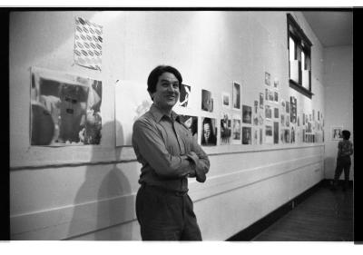  A medium-shot portrait of a person in a collared shirt with their arms crossed, addressing their attention to their right with raised eyebrows and a jovial expression. Behind them are pinned-up portraits on a gallery wall. 
