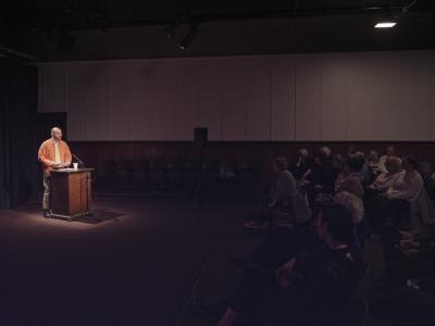A hall full of audience members face Cody Caetano while he speaks at a podium, under a spotlight. 
