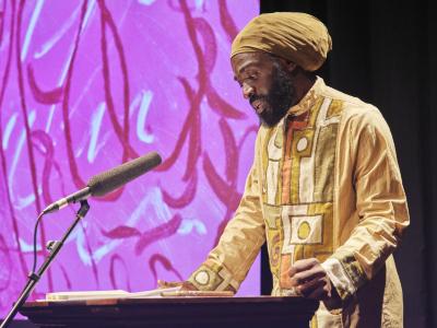 Phanuel Antwi stands behind a lectern and reads from a paper resting on its surface. He is adorned in traditional garments. To his right, there is a projection of Jonathan Alfaro’s pink, red, and white monotype print.