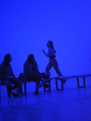 Nour Mobarak runs through a blue-lit gallery behind a row of audience members. The floor is marked with shapes made from neon tape. 