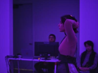 A hopeful look plasters Nour Mobarak’s face in this purple-lit side profile image. Her hands are clasped behind her head in what looks like a moment of pause. A technician at a desktop computer and one audience member is seen behind her.