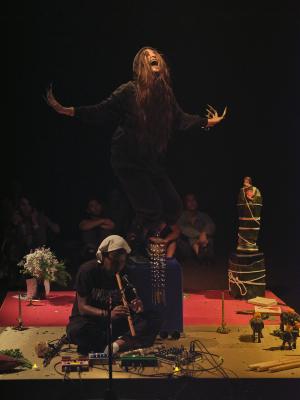 Tati sits cross legged on a square platform playing a wooden wind instrument. Bhenji stands on a blue box in the middle of said altar space, bending at the knees and devilishly bringing her hands toward the sky. She looks upward with a crazed look and an open mouth.