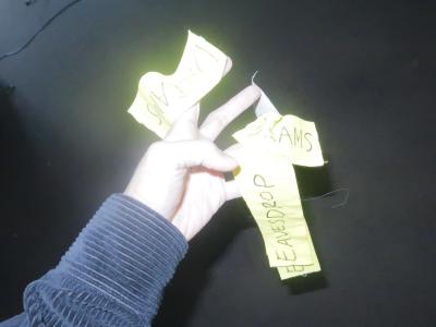 A hand reaches in front of the camera. The flash is bright and reflecting off of 3 yellow pieces of paper that stick to the person's fingers. One paper says DREAM, another says EAVESDROP, and the third one is crumpled with only the letters AMS visible. 