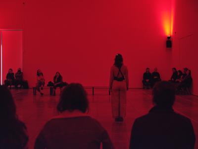 With her back to the camera, Nour Mobarak stands at the centre of a gallery space illuminated by a red glow. Her arms are tight at her sides and her feet together. Audience on all sides of her seated on benches.