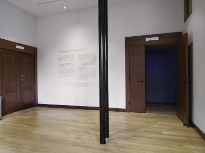 The entry foyer at The Western Front building. Two black poles stand in the middle of the room, the gallery door is open behind them, giving a glimpse of the exhibition's blue interior. Next to the gallery door is a wall with the exhibition's didactic text on it. 