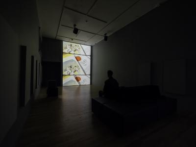 A visitor sits and watches an analogue video reel projected on a vertical wall in a dimly lit gallery. 