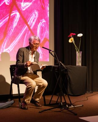A man with silver hair wearing a suit sits in a chair reading aloud from a book in front of a microphone. To his right there is a table covered by a black table cloth, and a vase with four flowers in it.