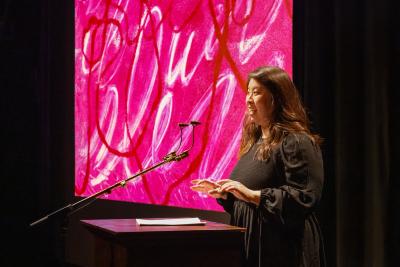 A woman wearing a black puff-sleeved dress is seen in profile at a wooden lectern. She energetically speaks into a microphone with her hands gesturing in front of her. A pink abstract image is projected in the background.