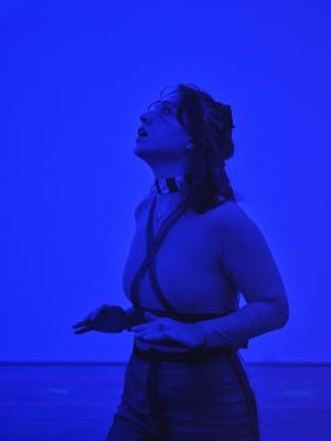 A portrait of Nour Mobarak in blue light. She looks up to the ceiling with her mouth agape. Her hair is disheveled from the movement of her performance, and she wears an x shaped harness over a long sleeve top. She keeps her arms above her waist, with her hands flat and palms facing down.