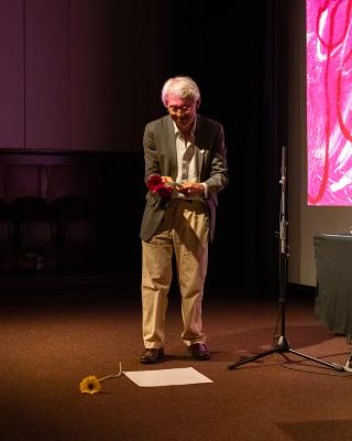 A man emotes a scrunched smile while tenderly holding a pink flower. A yellow flower and a blank piece of paper lay on the ground in front of him. A microphone is to his right.