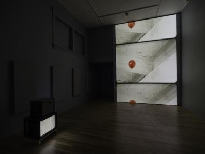 A large projection on a gallery wall shows a red balloon ascending in a closed space. On the floor, a small TV displays a video of the inside of a book, showing its pages with text..
