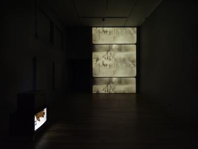 A large vertical projection of an analogue video reel on a gallery wall. A small television sits on the ground showing a video of a hand.