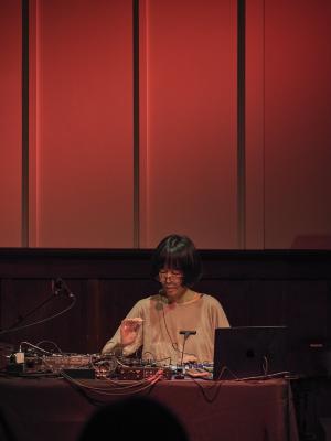 Phew stands behind a table covered by sound mixers, cables, and a laptop with one palm raised in the air with all fingers falling forward. A diffused red light shines above her head, highlighting the edges of the technology on the table. Her downwards gaze casts a green shadow of her headworn microphone onto her shoulder.