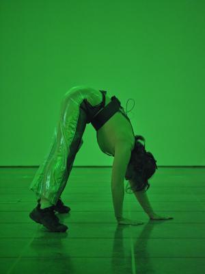 Green light fills the room as Nour Mobarak balances in a downward dog shape. She wears sneakers, metallic pants, and a harness with microphone wires. 