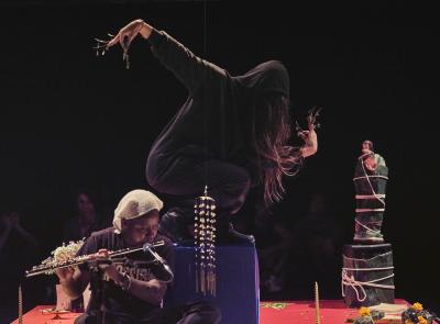 Tati au Miel passionately plays the flute in the lower left third of this picture. In the middle, Bhenji is pictured squatting atop the blue box. She has one arm extended behind her and the other bent at the elbow with her hand positioned in front of her face. She is wearing long gold fingernail jewelry.