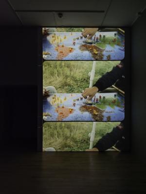 A large projection on a gallery wall shows two hands playing a board game on top of a world map.
