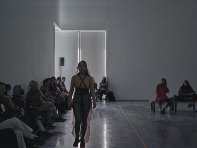 In a white cube gallery, Nour Mobarak walks towards the camera with her eyes closed. She wears an X shaped harness across her chest, with sheer fabric flowing behind her. Audience members sit against the walls, and coloured tape demarcates the performance area in x’s and lines on the floor.