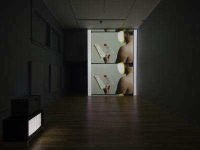 A large projection on a gallery wall shows the back of a person holding a book. The book features text and illustrations. On the floor, a small TV displays a video of the inside of the book, offering a close-up view of its pages.