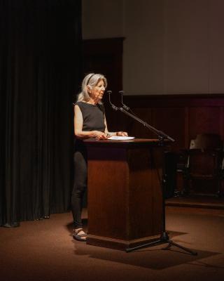 A woman with long gray hair reads off of a piece of paper into two small microphones at a wooden lectern. She wears all black and  is lit by a spotlight.