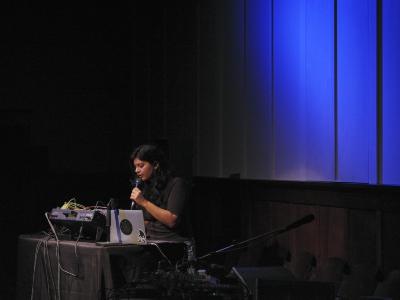 Piu vocalizes into a microphone from behind a laptop and sound mixer with multicoloured cables in the Grand Luxe Hall. A diffused blue light is cast onto the panelled white wall behind her while she is spotlit.