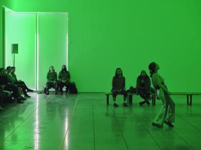 Green light fills a white-cube gallery space as Nour Mobarak performs in the centre of the room. She leans backwards and gazes up at the ceiling while her feet face forwards. Audience members sit on benches in the background, captivated by the performance. 