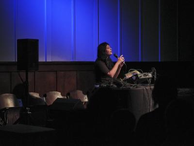 Piu vocalizes into a microphone with her eyes calmly closed and mouth relaxedly hung open. She is spotlit, with some of the light catching on the wooden theatre chairs of the Grand Luxe Hall folded up against the wall. The room is dark except for this leak of light and the diffused blue light is cast onto the panelled white wall behind her.