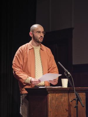 Cody Caetano stands at a wooden lectern, lit by a spotlight. He is in the middle of speech, gripping a stack of paper in his hands. He wears an orange jacket and a cream sweater. 