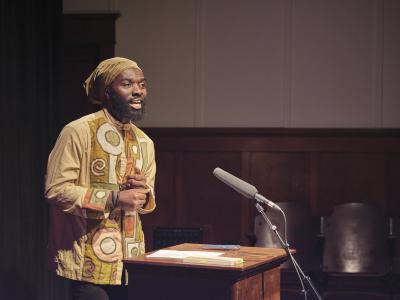 Phanuel Antwi speaks passionately with his hands clasped at his chest. He stands behind a lectern, speaking into a microphone. He is adorned in traditional garments.