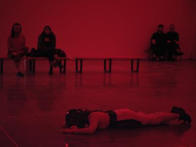Nour Mobarak lays face down on the floor with one arm tucked under her forehead. Her other arm is stretched in front of her, palm to the floor. The room is illuminated red and you can see four audience members seated on benches.