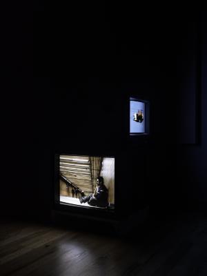 Two tv screens sit adjacent, one featuring a video of a man sitting on the ground, the other featuring a contraption descending from electrical wiring. 