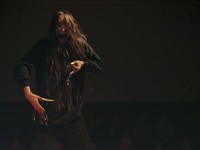 An energetic shot of Bhenji facing the camera, her mouth open as if she is screaming. She has black pigment on her teeth and is moving her hands in front of her, dangling her gold fingernail jewelry. She is looking down at her hands.
