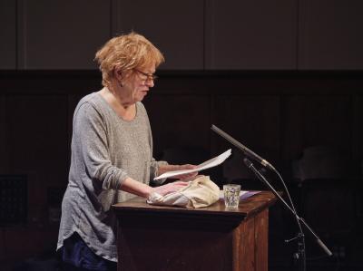 Dorothy Trujillo Lusk, stands at a podium, speaking to an audience. She wears a light grey sweater and brown glasses.  She grips a piece of paper with one hand while the other rests on a canvas bag atop the podium. 