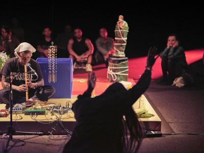 A focused Tati passionately plays a wooden wind instrument. The top half of Bhenji's body is silhouetted in the foreground, her hands raised above her head.