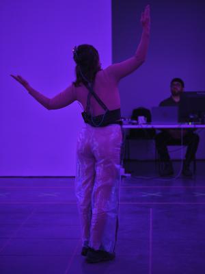 The gallery is flooded with purple light as Nour Mobarak stands with her back to the camera. She holds out her arms to create a U shape by bending upwards at the elbows. A technician sits at a desktop computer, focusing on the performance. 