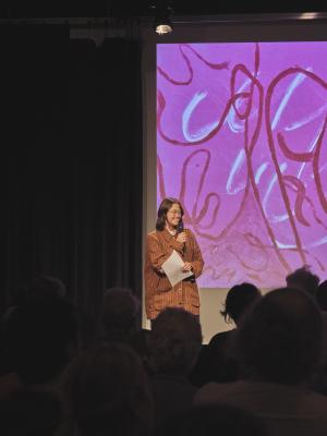 Kiel Torres stands holding a sheet of paper and speaking into a microphone before a crowd in the Grand Luxe Hall. A pink, red, and white monotype print by Jonathan Alfaro is projected behind her. 