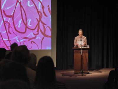 Tiziana La Melia stands behind a lectern. She smiles as she reads off a paper to a crowd that fills the Grand Luxe Hall. To her right, there is a projection of Jonathan Alfaro’s pink, red, and white monotype print.
