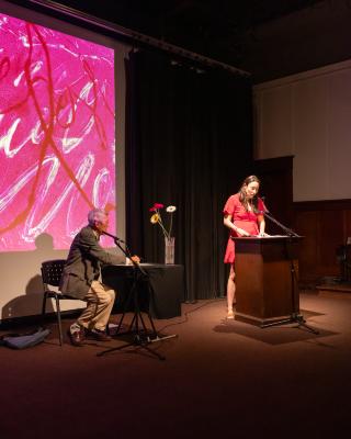 A woman wearing a red dress is spotlit at a wooden lectern as she reads into two small microphones. Meanwhile, a man wearing a suit sits behind her at a table covered by a black tablecloth with a vase of flowers on it. A pink abstract image is projected in the background. 