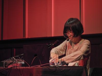 Phew stands behind a table covered by knobs, cables, and a laptop with her hands crossed over one another, touching the tabletop objects. She looks down at her hands through her glasses sitting at the tip of her nose. She is lit by a dim laptop screen and a soft red light that is diffused within the Grand Luxe Hall.